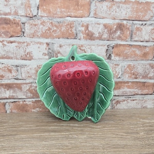 May include: A ceramic strawberry-shaped wall pocket with a glossy red finish. The strawberry is set on a green leaf-shaped base with detailed veining. The wall pocket has a hole at the top for hanging.