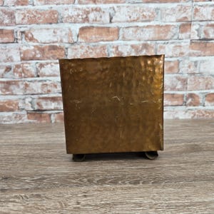 May include: A square-shaped container with a metallic bronze finish and a hammered texture. The object features a wavy top edge and is supported by four small, curved metal feet at the base, providing a stable foundation for the structure.