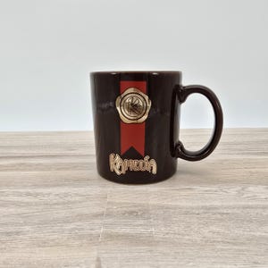 May include: A brown ceramic mug with a gold and red design. The mug features a gold seal with the letter "K" and the word "Kahlúa" in gold lettering.