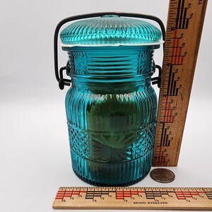 May include: A teal-colored glass jar with a ribbed design and a decorative lid. The jar has a metal handle and a white rubber seal. A wooden ruler is next to the jar. The jar is approximately 5 inches tall.