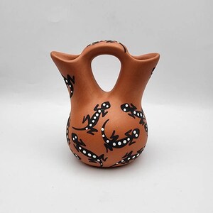 May include: A terracotta-colored pottery vase with a unique handle and two spouts. The vase is adorned with black and white lizard designs. The handmade vase is a decorative item.