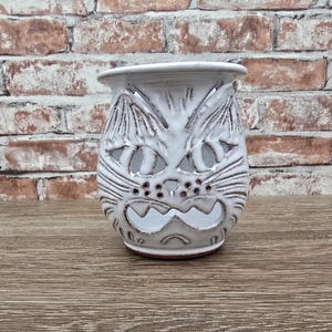 May include: White ceramic cat-shaped candle holder with carved details. The holder features a cat face design with cut-out eyes, nose, and mouth. The candle holder is on a wooden surface with a brick background.