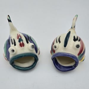 May include: Two ceramic fish-shaped containers with open mouths. Each piece features a cream-colored base with hand-painted vertical stripes in shades of purple, green, and red. The fish have prominent eyes and a single dorsal fin on top.