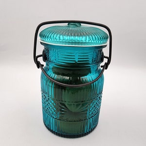 May include: A teal-colored glass jar with a lid and a black metal handle. The jar has a textured, ribbed design with a decorative pattern. The lid is also teal and has a ribbed design. The jar is likely used for storage or decoration.