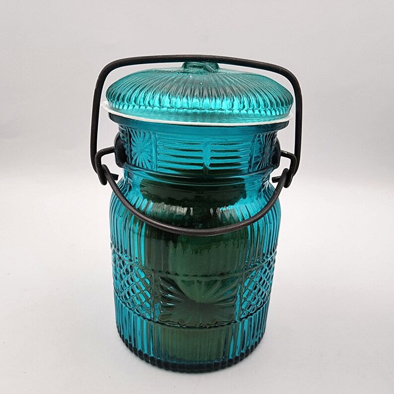 May include: A teal-colored glass jar with a lid and a black metal handle. The jar has a textured, ribbed design with a decorative pattern. The lid is also teal and has a ribbed design. The jar is likely used for storage or decoration.