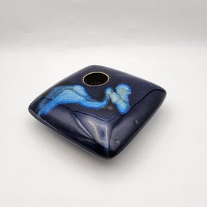 May include: A square, ceramic vase in a deep navy blue with a central opening. The vase features a flowing, abstract design in shades of blue and turquoise. The glossy finish enhances the colors.