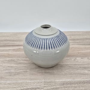 May include: A round, ceramic vase with a light gray body and a lid. The top portion of the vase features vertical blue stripes. The vase sits on a light brown wooden surface. The vase has a small opening at the top.