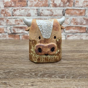 May include: A ceramic bull figurine featuring a textured tan and brown body with a white patch on the forehead. The bull has two curved white horns, dark circular eyes, and a prominent snout with two dark nostrils, crafted in a cylindrical shape.