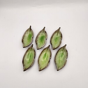 May include: A set of six small ceramic dishes shaped like leaves. Each dish features a glossy light green center with a darker green gradient and a brown, textured rim. The dishes are arranged in two rows of three, showcasing a smooth finish.