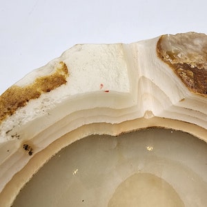 May include: A close-up of a natural stone ashtray. The ashtray is circular with a layered design, featuring shades of white, beige, and brown. The edges have a rough, natural texture, and the interior is smooth. The overall aesthetic is organic and earthy.