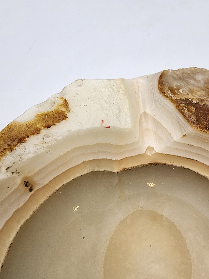 May include: A close-up of a natural stone ashtray. The ashtray is circular with a layered design, featuring shades of white, beige, and brown. The edges have a rough, natural texture, and the interior is smooth. The overall aesthetic is organic and earthy.