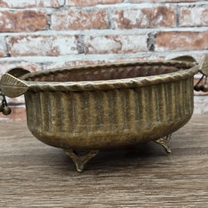 May include: Antique brass-toned oval planter with a ribbed body and three decorative feet. The rim has a twisted rope design, and leaf and berry accents adorn the sides. A vintage decorative piece.