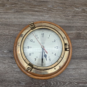 May include: A round quartz clock with a brass bezel and a wooden frame. The clock face is white with black numerals and hands. The clock has a nautical or maritime theme.