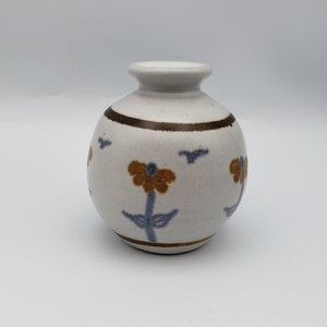 May include: A small, off-white ceramic vase with a round body and a narrow neck. The vase features brown and blue floral and bird designs. A brown band encircles the vase near the top. The vase is suitable for displaying small flowers or as a decorative item.