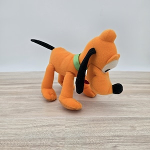 May include: Orange plush dog toy with black ears, tail, and nose. The dog has a green collar and a red tongue. The toy is standing on a light brown wooden surface.