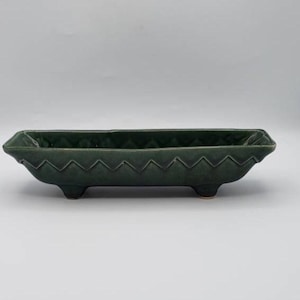 May include: A rectangular, dark forest green ceramic planter with a distinct zigzag or chevron pattern along the upper rim. The vessel features a smooth, glazed finish and is supported by four small, rounded feet at the base, creating a low profile.