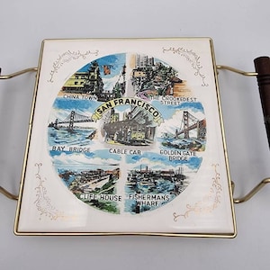 May include: A white ceramic serving tray with a gold trim and a colorful illustration of San Francisco landmarks. The tray features the Golden Gate Bridge, Fisherman's Wharf, Cable Car, and more. The tray has a handle on each side.