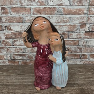 May include: A ceramic figurine depicting two figures, one larger and one smaller, both with stylized features. The larger figure wears a burgundy dress and a black hood, while the smaller figure wears a blue and white striped dress. The figures are standing close together.