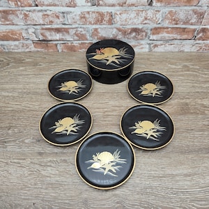 May include: A black lacquer coaster set with a matching round box. Each coaster and the box lid feature a gold design of a sun, shells, and seaweed. The coasters have gold rims and are arranged around the box.