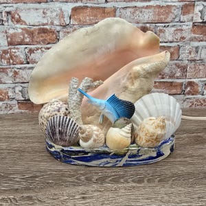 May include: A decorative display featuring a large cream-colored conch shell, a blue sailfish figurine, white coral, and various smaller seashells arranged on a circular base with a blue and white marbled pattern, creating a coastal arrangement.