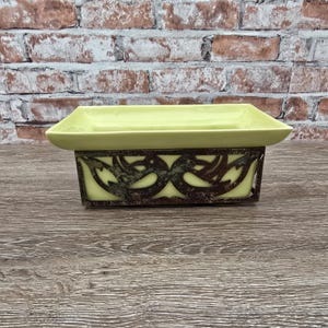 May include: A rectangular light yellow ceramic dish set within a dark brown metal frame. The metal frame features a decorative cutout pattern of two mirrored bird-like shapes. The dish has a smooth, wide rim and a deep, flat-bottomed basin.