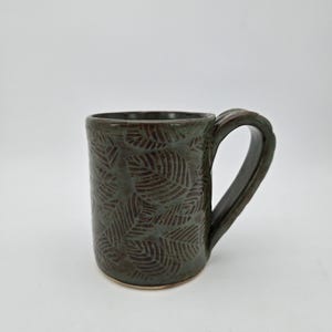 May include: A dark green ceramic mug with a leaf pattern. The mug has a curved handle and a textured surface. The mug is approximately 4 inches tall and 3 inches in diameter, perfect for coffee or tea.