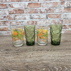 May include: Four vintage drinking glasses. Two clear glasses with orange and yellow floral designs, and two textured green glasses. The glasses are arranged on a wooden surface with a brick background.