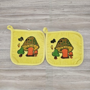 May include: A set of two yellow pot holders with a mushroom design. The mushrooms are brown with orange stems and green leaves. The pot holders have a loop for hanging.