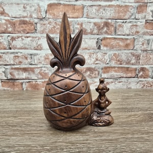 May include: A brown, carved wooden pineapple figurine with a small figure seated beside it. The pineapple has a textured surface with diamond-shaped patterns and a spiky top. The small figure is wearing a necklace.