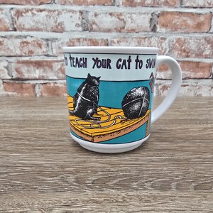 May include: A white ceramic mug featuring a graphic illustration of a black cat sitting on a yellow wooden dock next to a ball of yarn. The text printed on the mug reads, "TO TEACH YOUR CAT TO SWIM". The mug has a curved handle and a smooth finish.