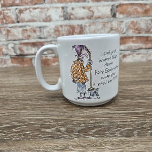 May include: A white ceramic mug featuring a cartoon illustration of a person in a checkered shirt and nightcap holding a mop and bucket. The text reads: ...and just where's that damn Fairy Godmother when you need her????