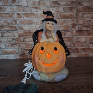May include: A ceramic Halloween lamp featuring a witch with gray hair and a black hat resting her chin on her hand. She is positioned behind a large, orange, carved jack-o'-lantern with a glowing face. The base is textured and gray.