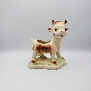 May include: A ceramic cow-shaped planter featuring a cream-colored body with brown shading on the sides and face. The cow has pink cheeks, dark eyes, and small horns, standing on a light green base with a hollow opening on its back for plants.