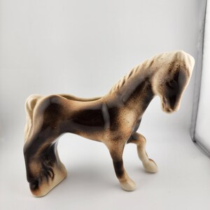 May include: A ceramic horse figurine in shades of brown and tan. The horse is posed in a walking stance, with its head lowered. The figurine has a hollowed-out back, suggesting it could be used as a planter or decorative piece.