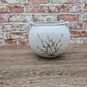 May include: A white ceramic lidded jar with a round body and two small handles. The jar features a floral design with pink flowers and green leaves. The lid has a twisted handle. The jar is on a wooden surface in front of a brick wall.