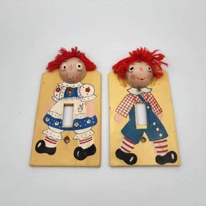 May include: Two vintage, hand-painted light switch covers. Each features a doll design with red yarn hair, a wooden head, and a painted outfit. One doll wears a blue and white dress, the other a blue jumpsuit. The background is yellow.