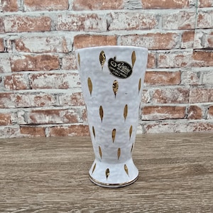 May include: White ceramic vase with gold leaf accents and a flared base. The vase has a label that reads "Rossini Originals Hand Decorated Italy". The vase is approximately 8 inches tall.