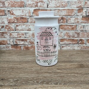 May include: A white ceramic jar with a lid, featuring a hand-drawn illustration of a house, a person, and the text "For my home." The design includes pink accents and black outlines, set against a brick background.