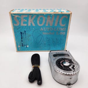 May include: A silver Sekonic Auto-Lumi model L-86 light meter with a black dial, a black neck strap, and its original blue and white cardboard box featuring the brand name and model number printed in large silver lettering on the front panel.