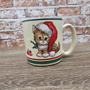 May include: Cream-colored ceramic mug featuring a cartoon kitten wearing a Santa hat. The mug has a green rim and a red and green stripe near the base. A red mouse is also depicted on the mug. The handle is off-white.