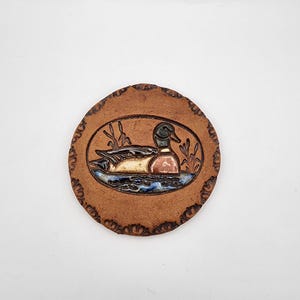 May include: A circular terracotta-colored ceramic coaster featuring a central oval relief of a mallard duck swimming in water among reeds. The outer edge of the coaster is decorated with a repeating stamped pattern in a dark brown glaze.
