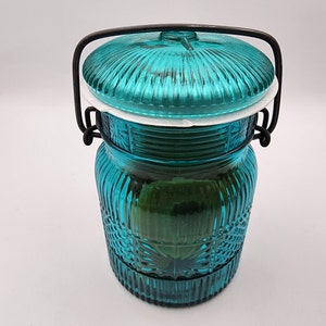May include: A teal glass jar with a lid and a black metal handle. The jar has a ribbed design on the lid and body, with a decorative pattern near the base. The jar is approximately 7 inches tall.