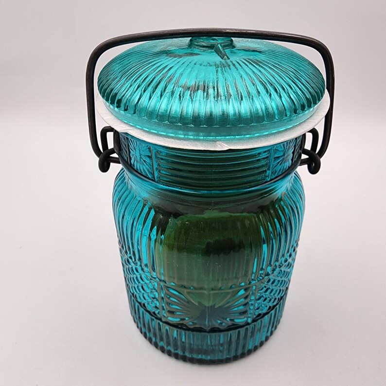 May include: A teal glass jar with a lid and a black metal handle. The jar has a ribbed design on the lid and body, with a decorative pattern near the base. The jar is approximately 7 inches tall.