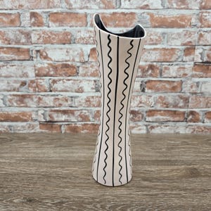 May include: A tall, slender ceramic vase with a cream-colored exterior featuring vertical black wavy lines. The vase has a distinctively curved, asymmetrical top rim and a smooth, glossy black interior finish that contrasts with the light exterior.