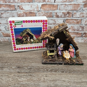 May include: A 4-piece Italian nativity set featuring a wooden stable structure covered in moss, with hand-painted figurines of a person in purple, a person in pink and blue, and a baby in a manger, accompanied by its original cardboard box.