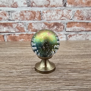 May include: A decorative glass orb with a brass base. The orb features a blend of green, yellow, and blue hues with a bubble-like texture. The brass base has a classic, elegant design, adding to the overall aesthetic.