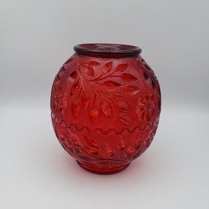 May include: A red glass vase with an oval shape and intricate floral and leaf patterns. The vase has a wide opening and a textured surface. The design includes detailed embossed flowers and foliage, creating a decorative and elegant appearance.