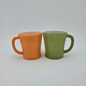 May include: Two glass mugs with a smooth, opaque finish. One mug is a vibrant orange color, and the other is a soft, muted green. Both mugs feature a rounded handle on the side and a slightly flared rim, standing side by side against a plain background.