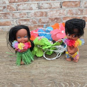 May include: Two vintage Hawaiian hula dolls with black hair, flower leis, and grass skirts. One doll is wearing a green grass skirt, and the other is wearing floral shorts. A string of colorful flower lights is between the dolls.