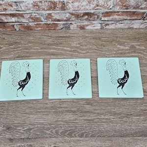 May include: Three square coasters with a light teal background. Each coaster features a black and white illustration of a rooster. The rooster is standing with its head up and its tail feathers fanned out. The coasters are arranged on a wooden surface.